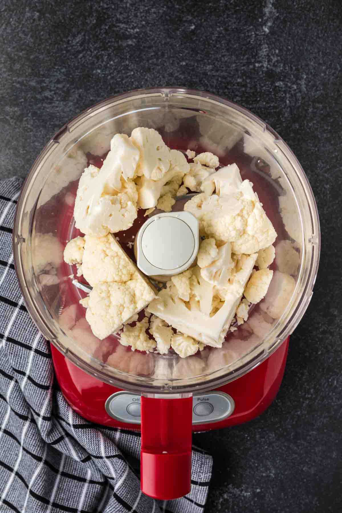 A red food processor filled with cauliflower florets sits on a dark countertop, ready to transform into perfectly seasoned cauliflower rice. A black and white checkered cloth is placed beside it. The transparent lid reveals the fresh cauliflower inside, inviting culinary creativity.