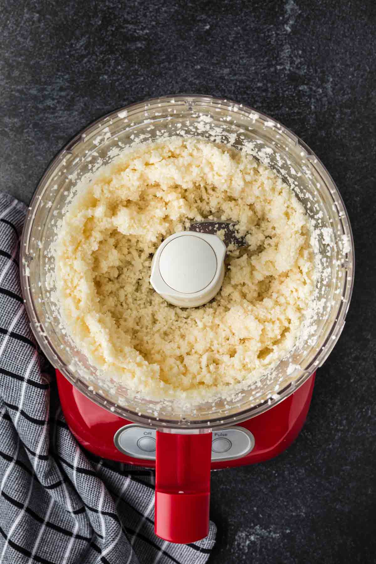 A red food processor containing finely chopped cauliflower rice sits on a dark countertop, ready for seasoning. Beside it, a gray and white checkered cloth adds a subtle touch of charm.