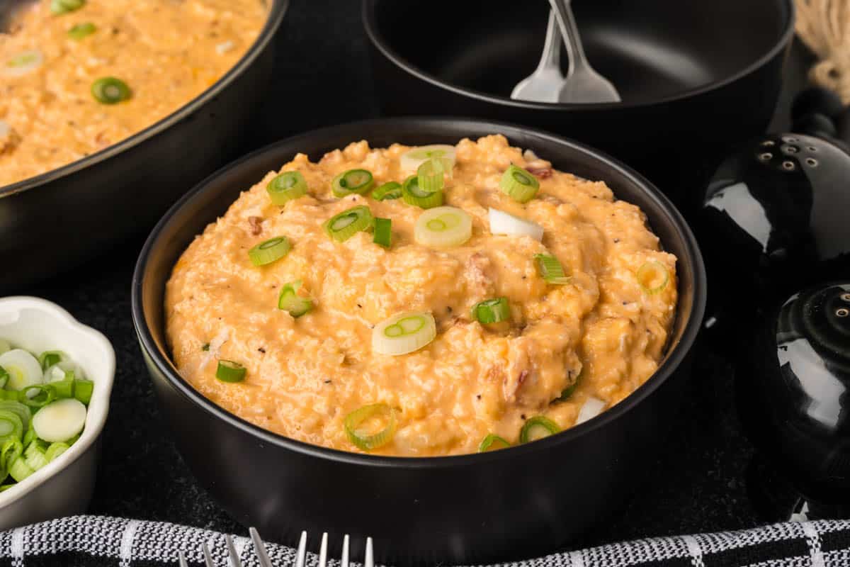 A bowl of creamy potato salad topped with sliced green onions, reminiscent of a perfectly seasoned dish. Served in a black bowl, it sits alongside similar dishes, with a nod to how to season cauliflower rice, and a small bowl of green onions in the background adds an extra touch.