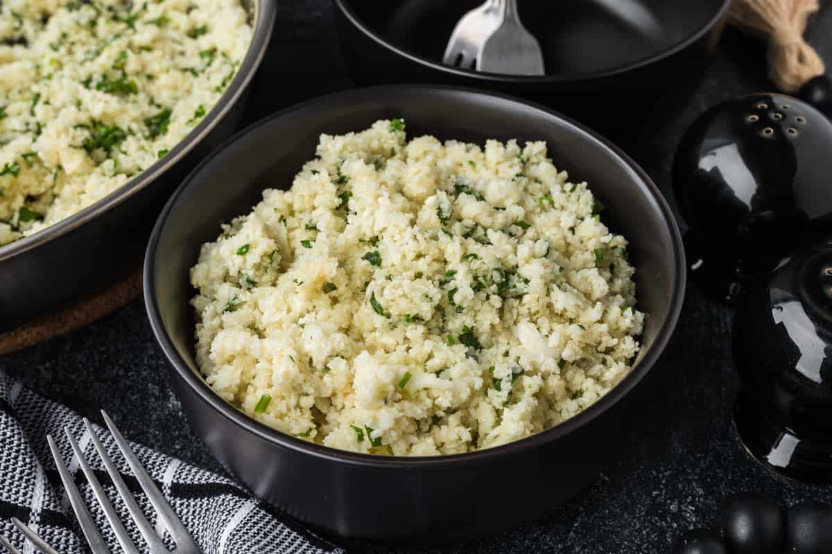 A bowl of cauliflower rice, seasoned to perfection with herbs, sits on a dark surface. A fork rests on a black plate nearby, alongside a black pepper grinder that enhances the kitchen setting. In the foreground is a folded striped cloth, completing this guide on how to season cauliflower rice elegantly.