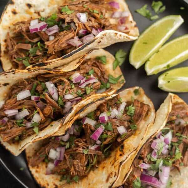 Four tacos filled with slow cooker barbacoa, shredded meat, chopped red onions, and cilantro are arranged on a dark plate. Lime wedges are placed on the side. The tacos are served in lightly charred tortillas.