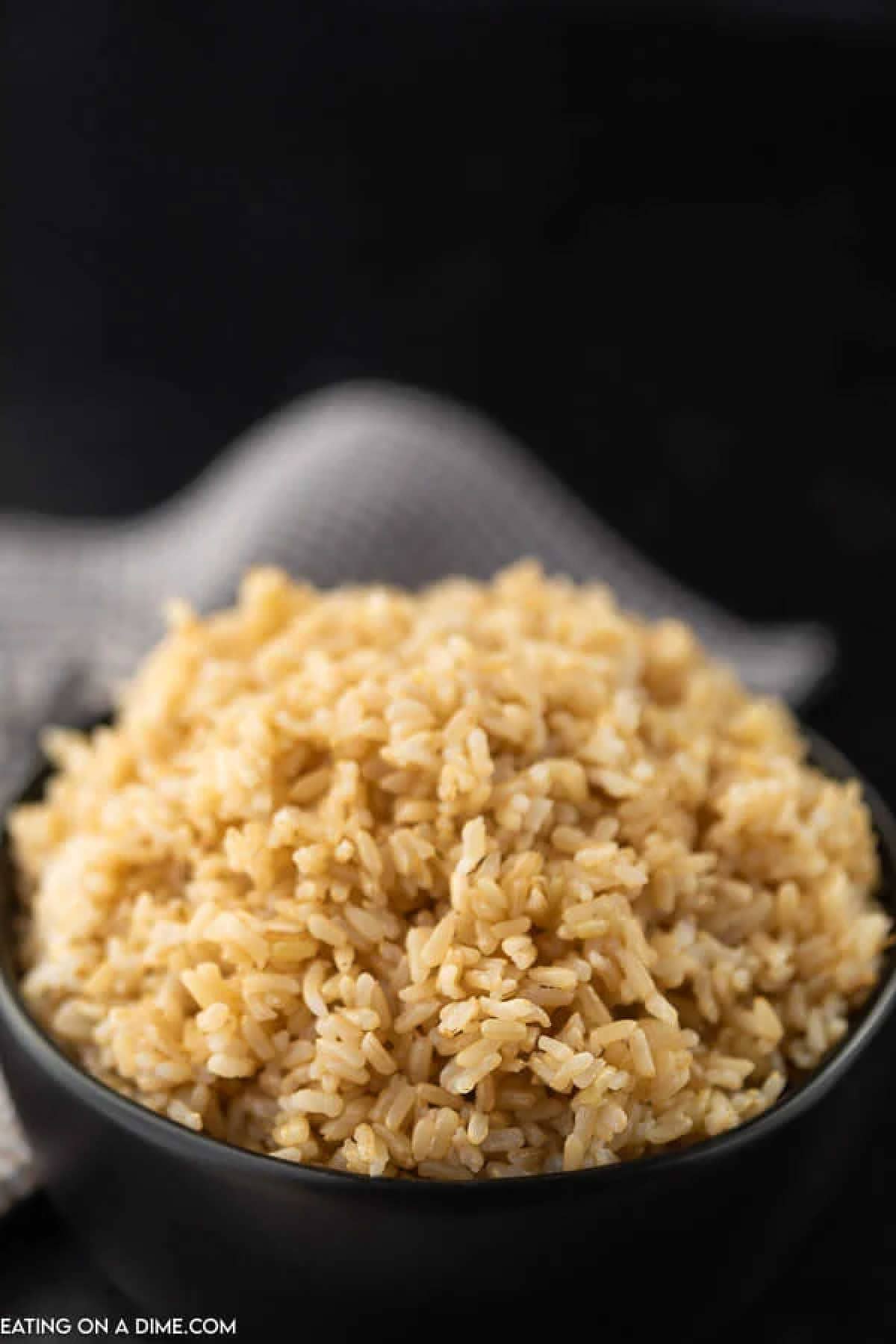 A bowl filled with fluffy, cooked brown rice sits against a dark background. A textured gray cloth peeks from behind, adding depth. The rice appears light and airy, filling the bowl to the brim, inviting you to explore how to season brown rice perfectly.