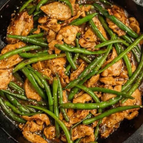 A close-up of a stir-fry dish in a skillet, featuring sliced chicken pieces and green beans cooked in a savory sauce reminiscent of Panda Express String Bean Chicken. The dish is garnished with sesame seeds, giving it a flavorful and visually appealing presentation.