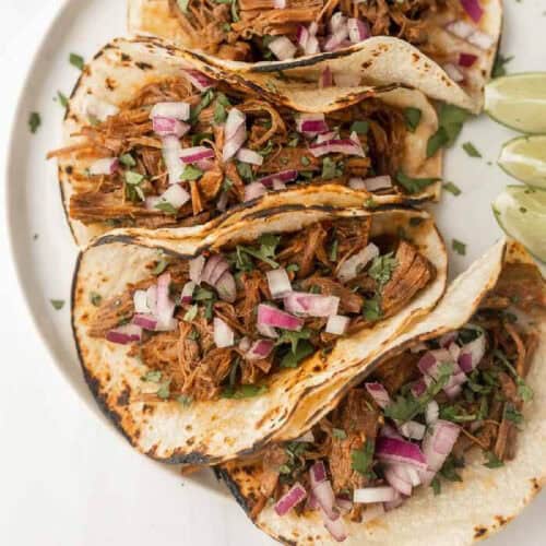 Three tacos filled with shredded barbacoa beef, chopped red onions, and cilantro are arranged on a white plate. The tacos, crafted from an Instant Pot recipe, are topped with lime wedges on the side. The tortillas appear slightly charred and toasted.