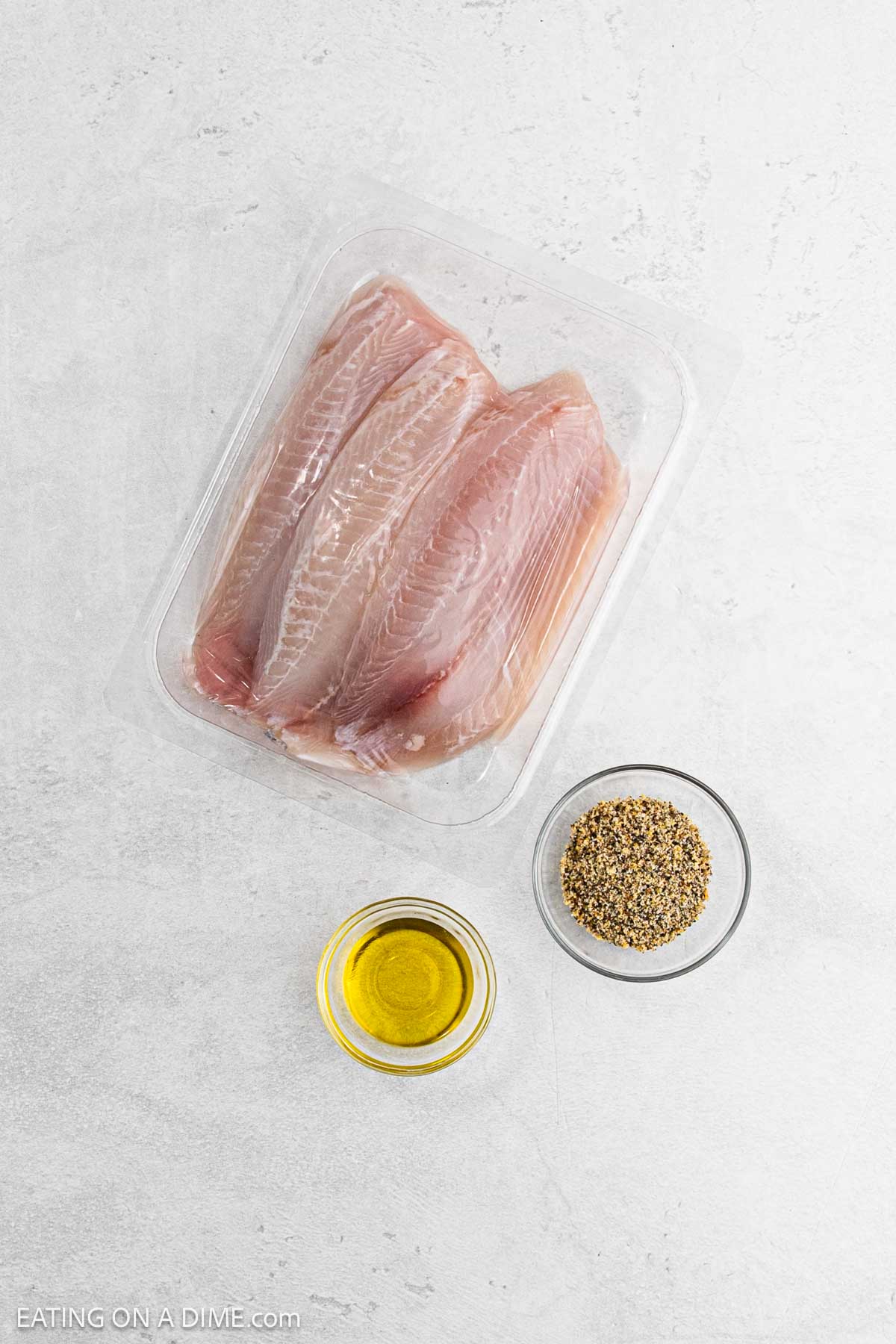 A plastic tray with raw tilapia fillets rests on a gray surface, ready to become delicious grilled tilapia. Next to it are a small bowl of oil and a bowl filled with a savory seasoning mix.