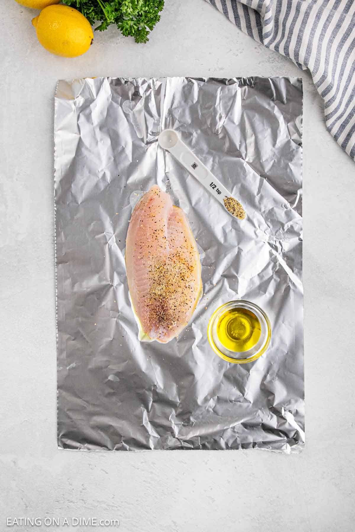 A raw, seasoned chicken breast lies on aluminum foil beside a small, clear glass bowl of olive oil and a teaspoon with spices. A striped cloth, lemons, and parsley complete the fresh scene reminiscent of preparing grilled tilapia.