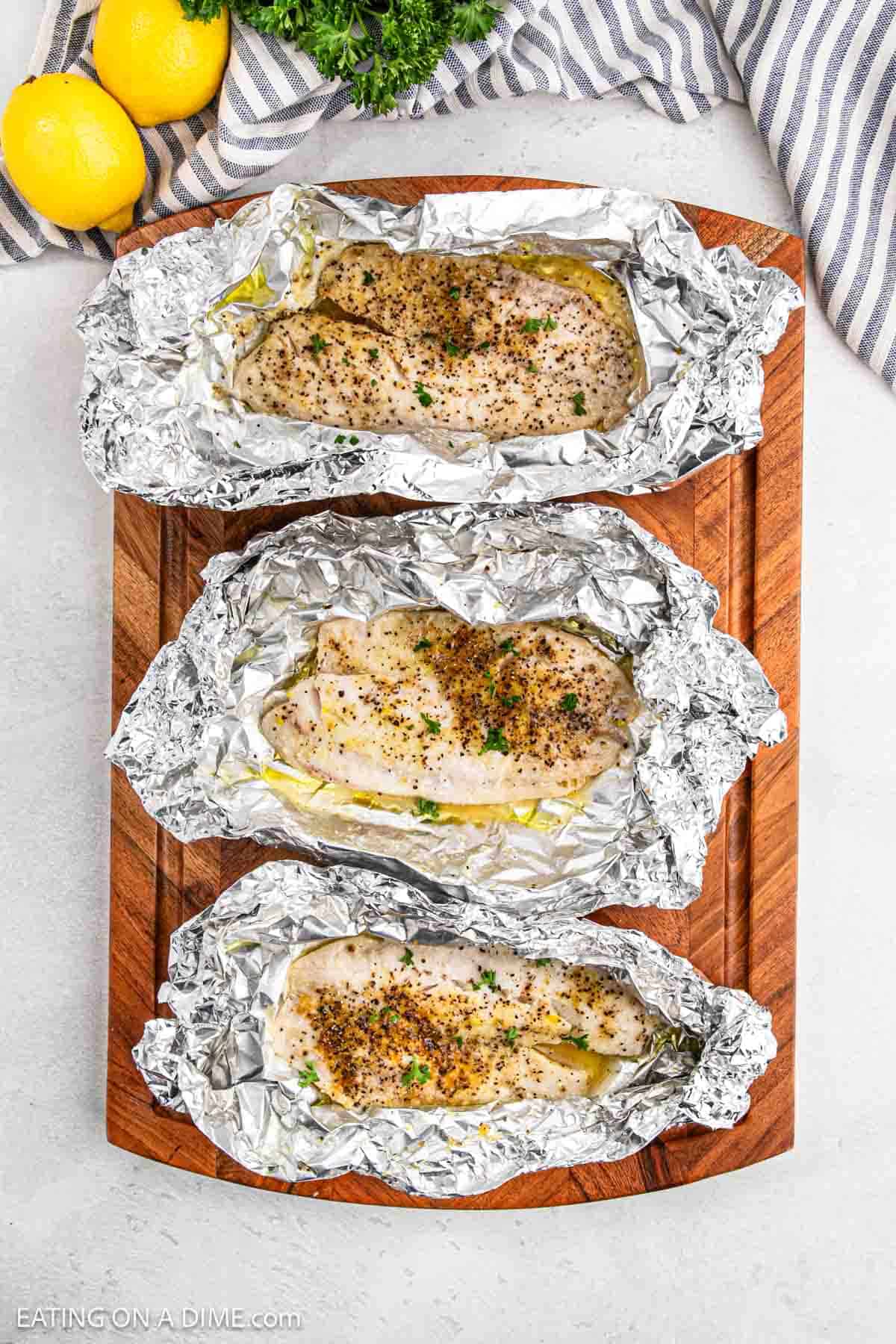 Three seasoned chicken breasts are cooked alongside grilled tilapia in foil packets on a wooden board. Fresh parsley is sprinkled on top. Lemons and a striped fabric are in the background.