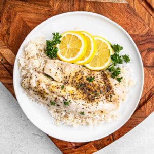 A plate with a seasoned grilled tilapia fillet on a bed of white rice, garnished with slices of lemon and parsley. The plate rests on a wooden surface.