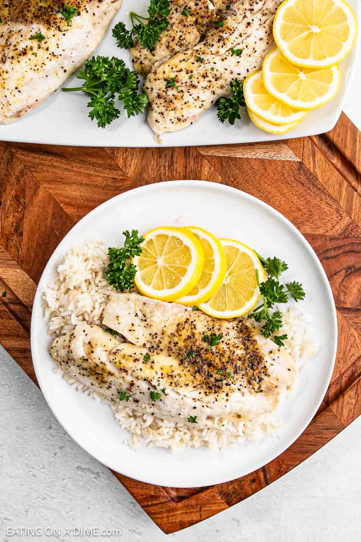 A plate of grilled tilapia fillets on a bed of white rice, garnished with lemon slices and parsley. Another plate with more fillets, lemon, and parsley is visible in the background on a wooden surface.