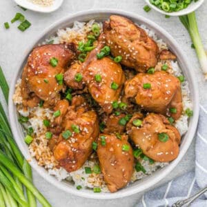 A bowl of crock pot honey garlic chicken thighs, glazed to perfection and garnished with sesame seeds and chopped green onions, rests on a bed of rice. Green onions and a spoon lie nearby on a light-colored surface.