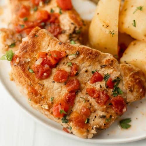 A plate with grilled pork chops topped with diced tomatoes and herbs, reminiscent of an Italian recipe, served alongside seasoned potato wedges. The pork chops have a golden-brown crust, and the dish is garnished with parsley.