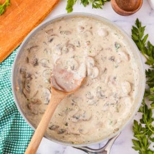 A saucepan filled with creamy mushroom sauce, with slices of mushrooms visible, being stirred by a wooden spoon. The pan is placed on a white marble countertop, surrounded by fresh parsley leaves, a green checkered cloth, a pepper mill, and a cutting board. Discover this delightful creamy mushroom sauce recipe today!