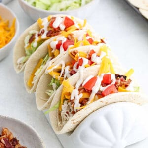 Three Chicken Bacon Ranch Tacos feature shredded cheese, diced tomatoes, crispy bacon, and lettuce drizzled with a creamy ranch sauce, all nestled in a taco holder. Additional bowls of cheese and toppings are visible in the background.