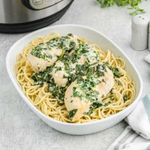 A white dish filled with an Instant Pot Chicken Florentine recipe features cooked spaghetti topped with creamy spinach and chicken breasts. Behind it, an instant pot and a salt shaker grace the kitchen countertop, while a cloth napkin is neatly placed beside the dish.