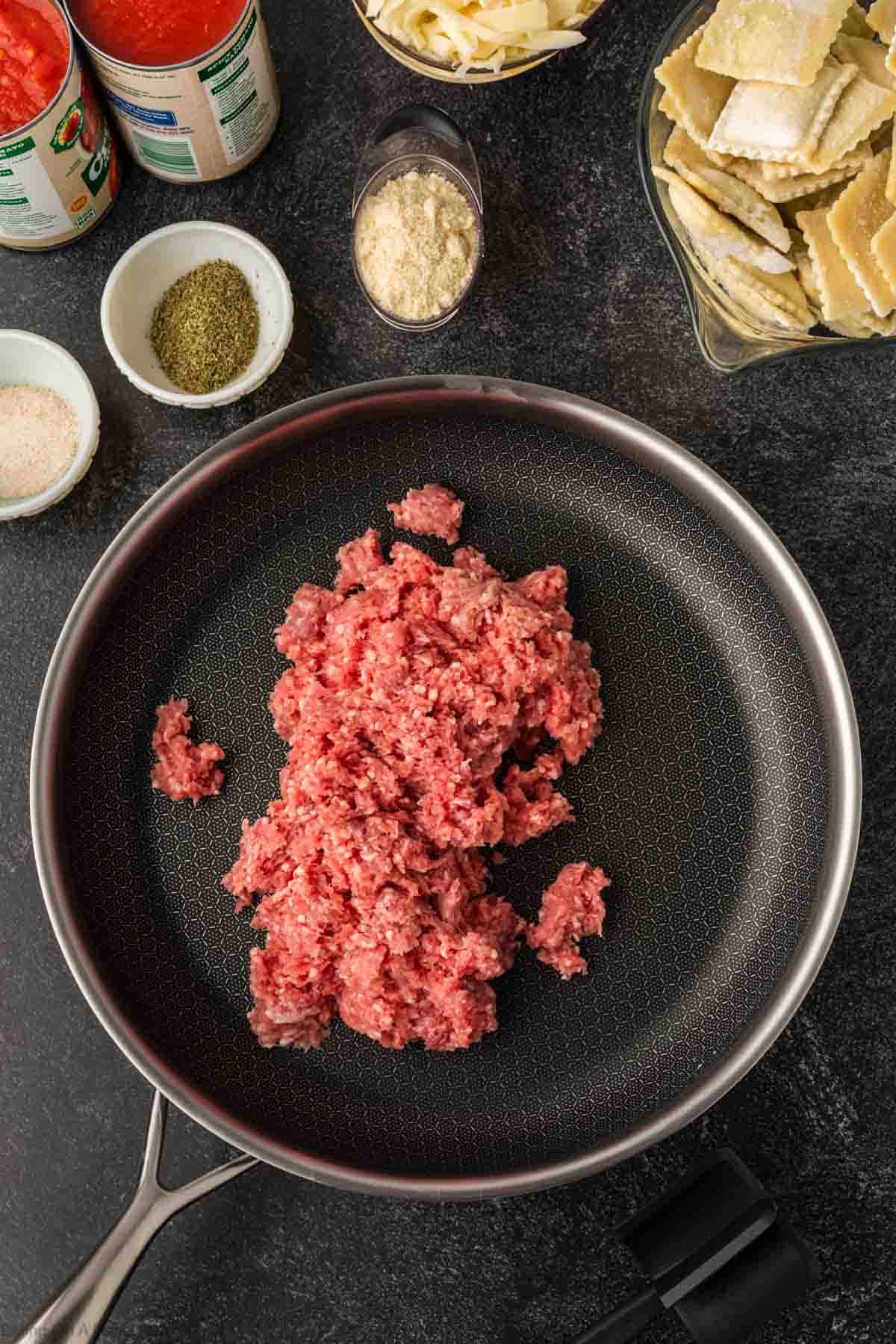 Ground Beef in a skillet with a bowl of frozen ravioli on the side with small bowls of seasoning and cheese