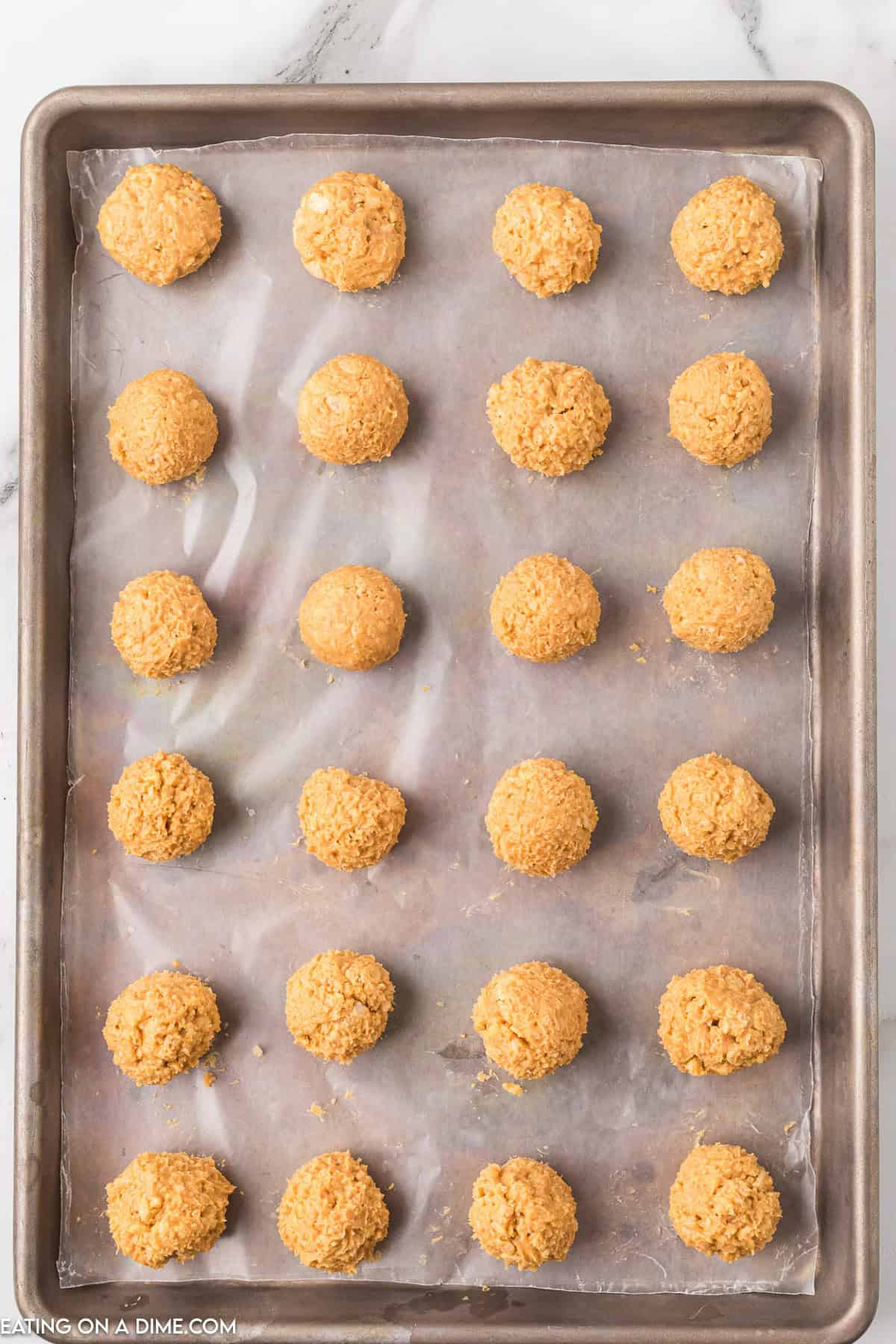 A baking tray lined with parchment paper holds 20 evenly spaced, round, uncooked sausage balls. The balls are a light golden color and placed in a grid pattern ready to be baked. The surface beneath the tray is white marble, reminiscent of preparing Chocolate Peanut Butter Balls with Rice Krispies.