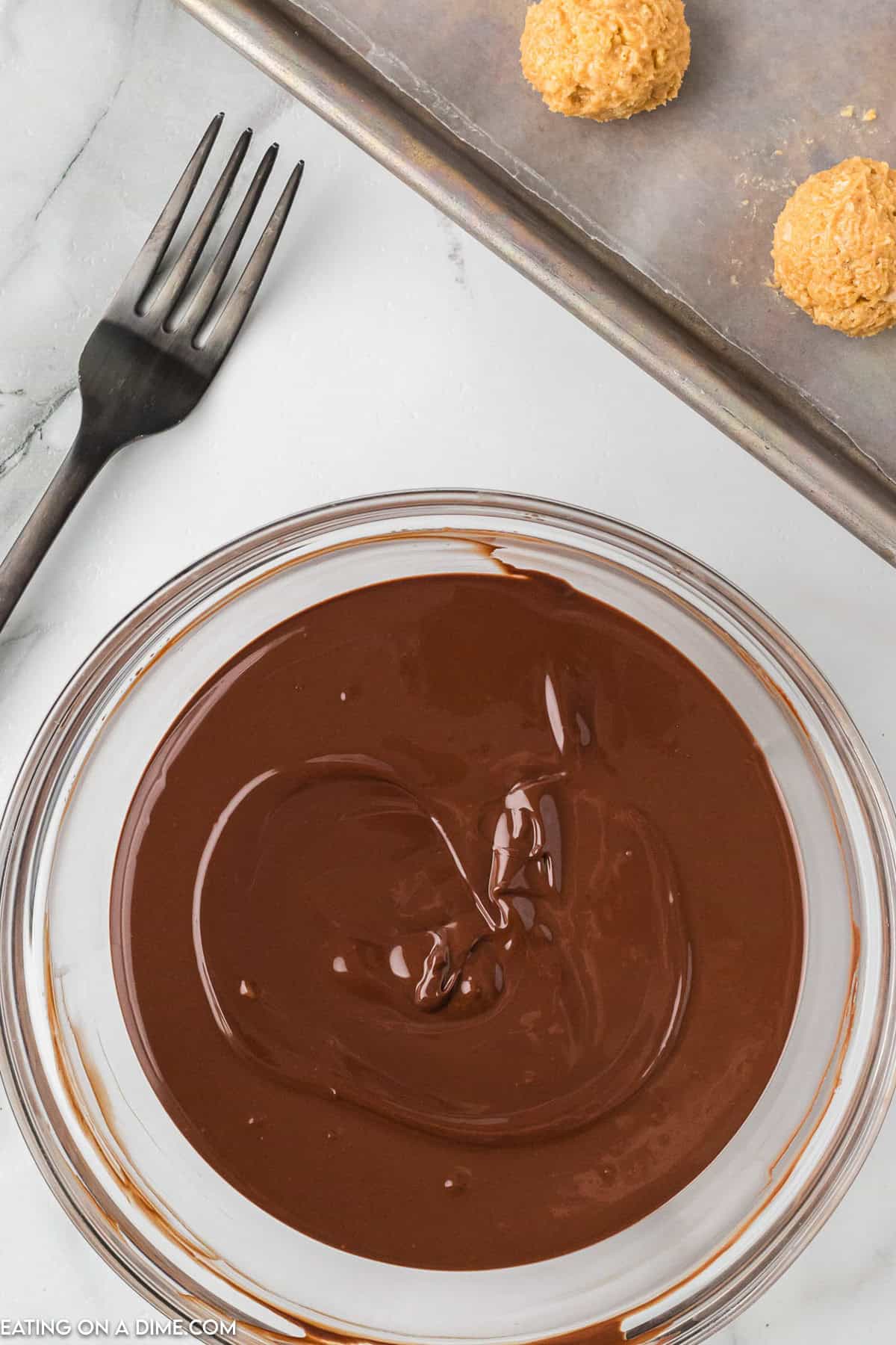 A glass bowl filled with melted chocolate is placed on a marble surface. Next to it is a black fork and a baking tray with parchment paper holding two uncoated Chocolate Peanut Butter Balls with Rice Krispies. The scene suggests preparation for dipping the treats in the chocolate.