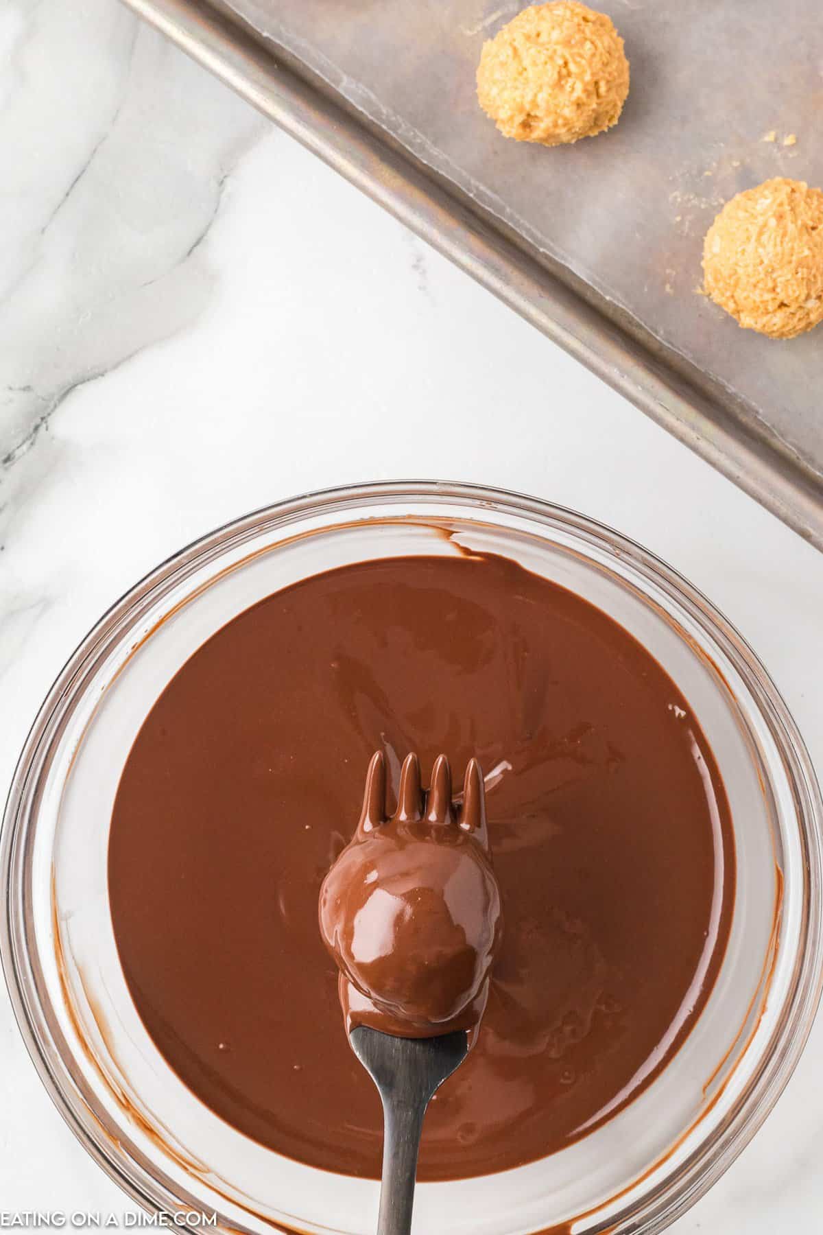 A fork holds a ball of confection dipped in smooth melted chocolate above a glass bowl. In the background, a baking sheet with wax paper holds two undipped Chocolate Peanut Butter Balls with Rice Krispies, contrasting against the marble countertop.