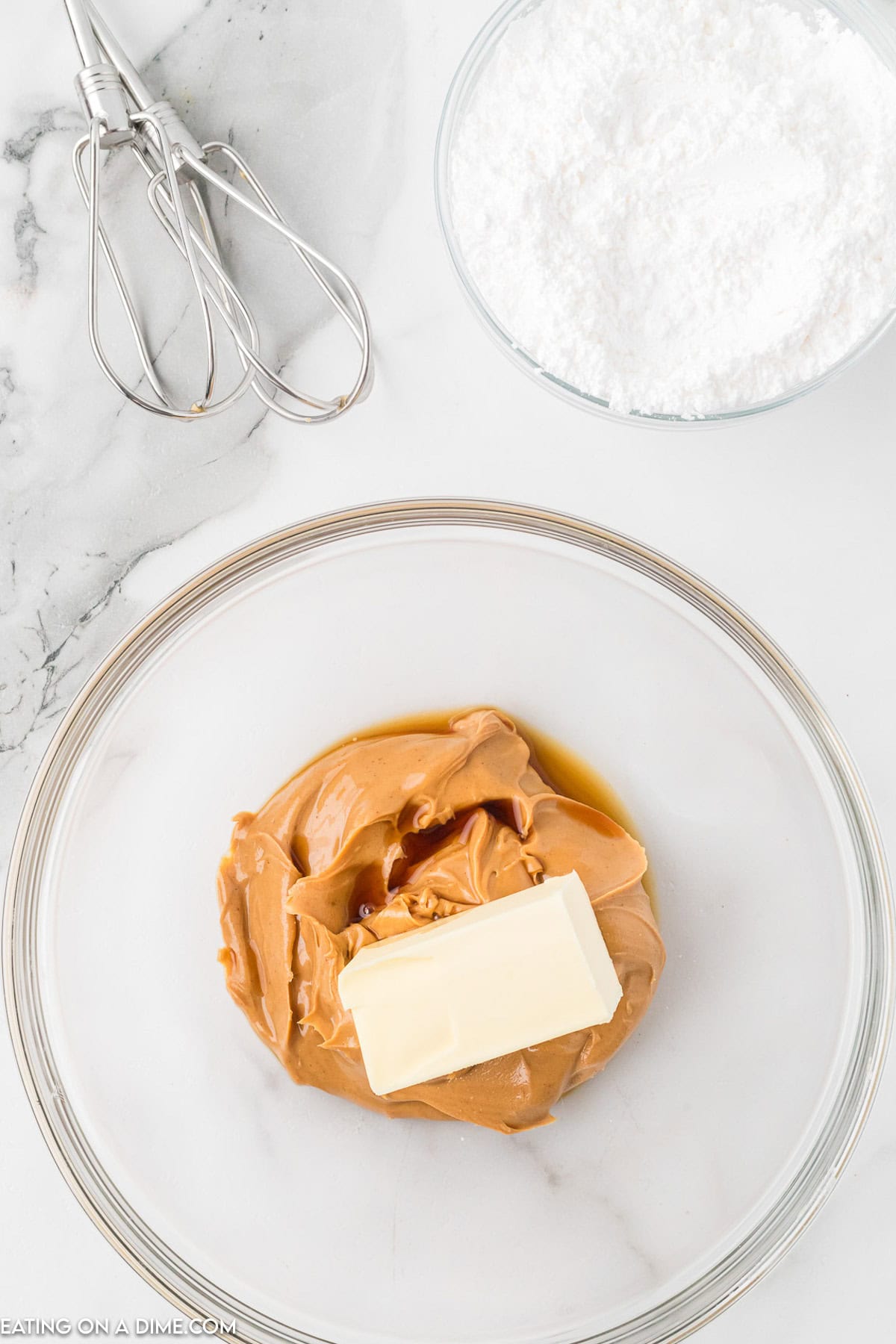 A glass mixing bowl contains a large dollop of peanut butter and a stick of butter on a marble countertop, ready to make Chocolate Peanut Butter Balls with Rice Krispies. Nearby, a small bowl brims with powdered sugar while a hand mixer with beaters rests on the counter.