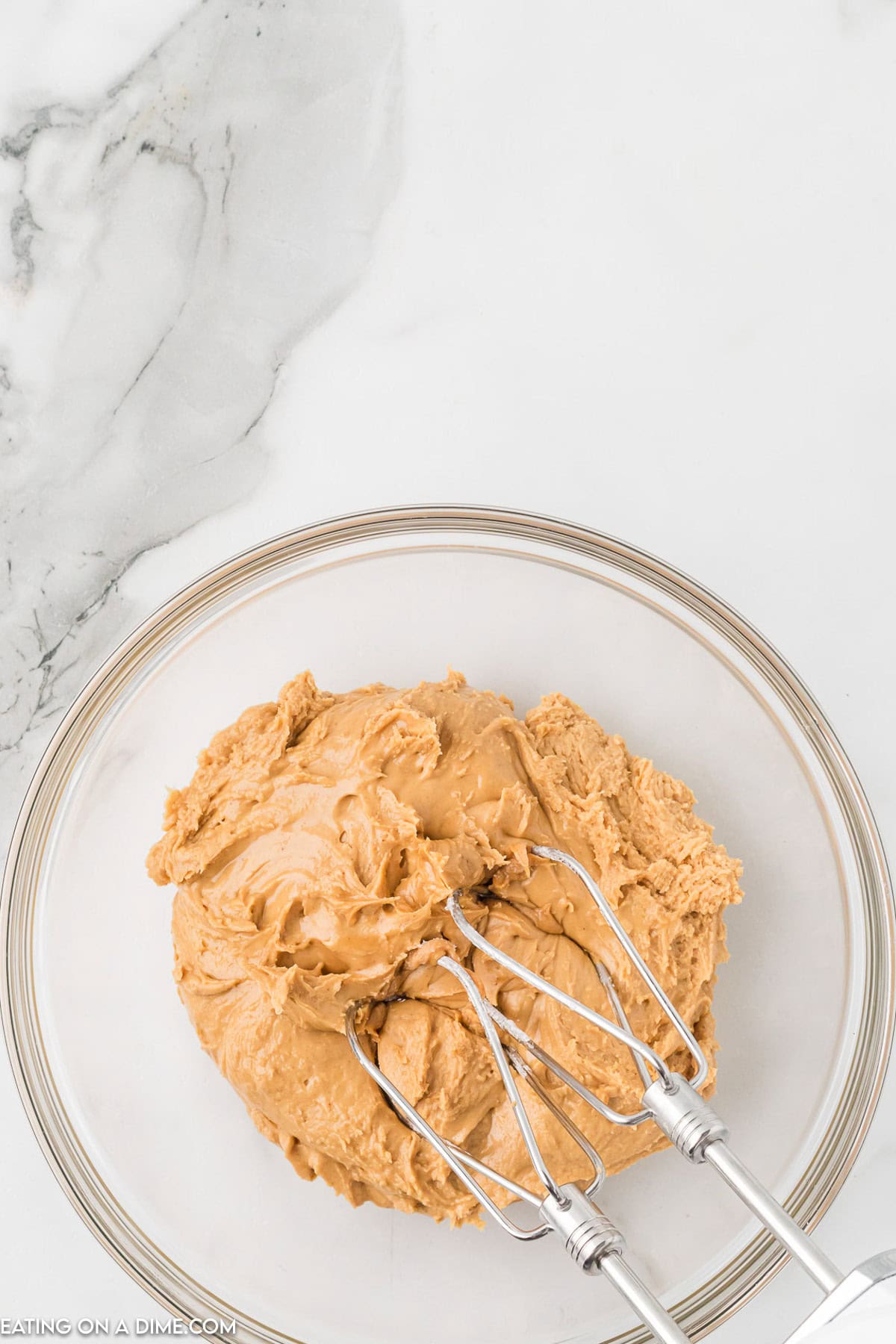 A glass mixing bowl filled with a creamy peanut butter mixture for Chocolate Peanut Butter Balls with Rice Krispies is being mixed with an electric hand mixer on a marble countertop.