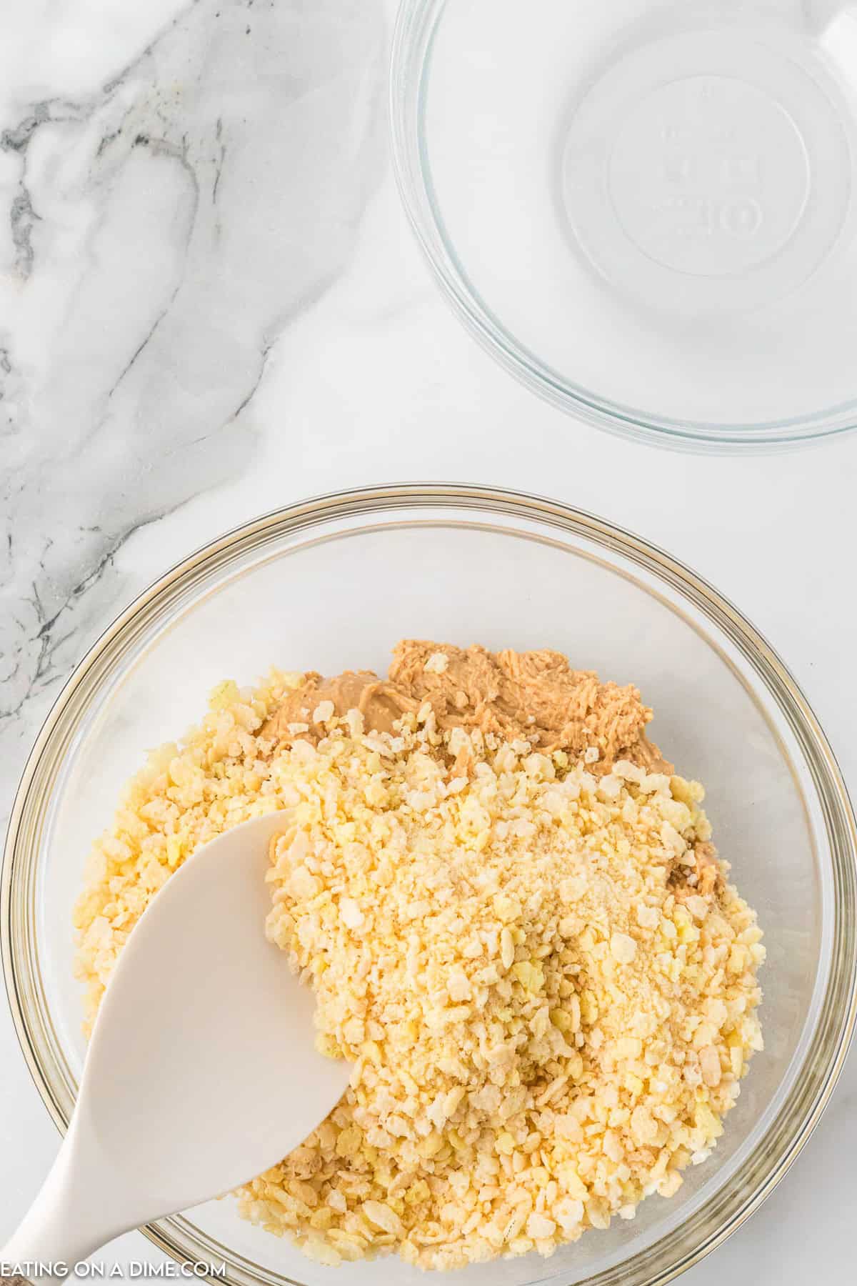 A glass bowl containing a mixture of crushed rice cereal and cookie butter is being stirred with a white spatula, reminiscent of preparing Chocolate Peanut Butter Balls with Rice Krispies. The ingredients rest on a marble countertop, with another empty glass bowl visible in the upper right corner.