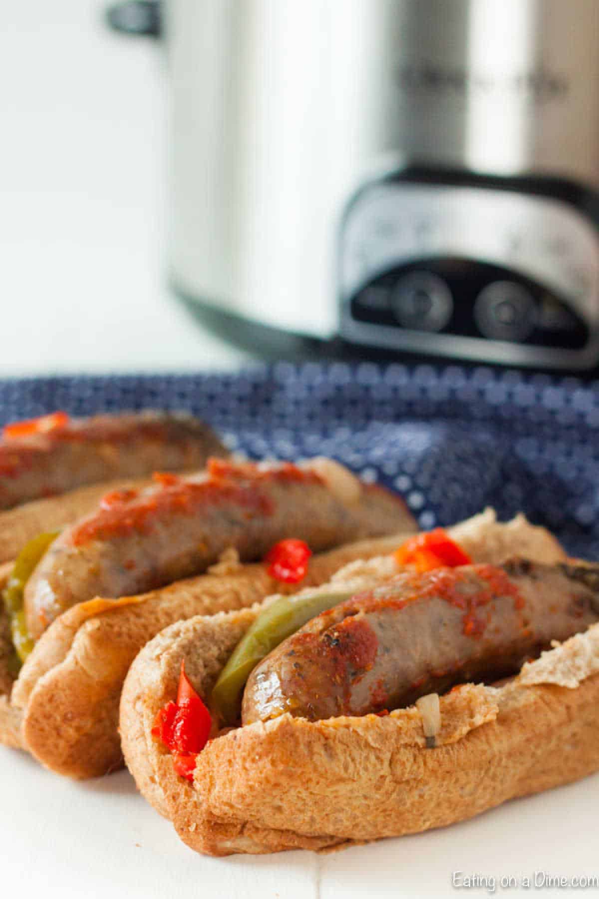 Three grilled sausages topped with red and green peppers rest in open buns in front of the Crockpot, simmering the ultimate sausage and peppers delight. A blue cloth is partially visible underneath.