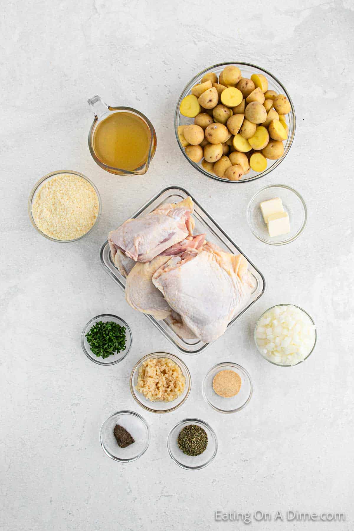 Top-view image of various ingredients laid out on a white surface, perfect for making Slow Cooker Garlic Parmesan Chicken. Includes bone-in chicken thighs, halved baby potatoes, a cup of broth, a block of butter, diced onions, minced garlic, chopped parsley, grated parmesan, and various spices in small bowls.