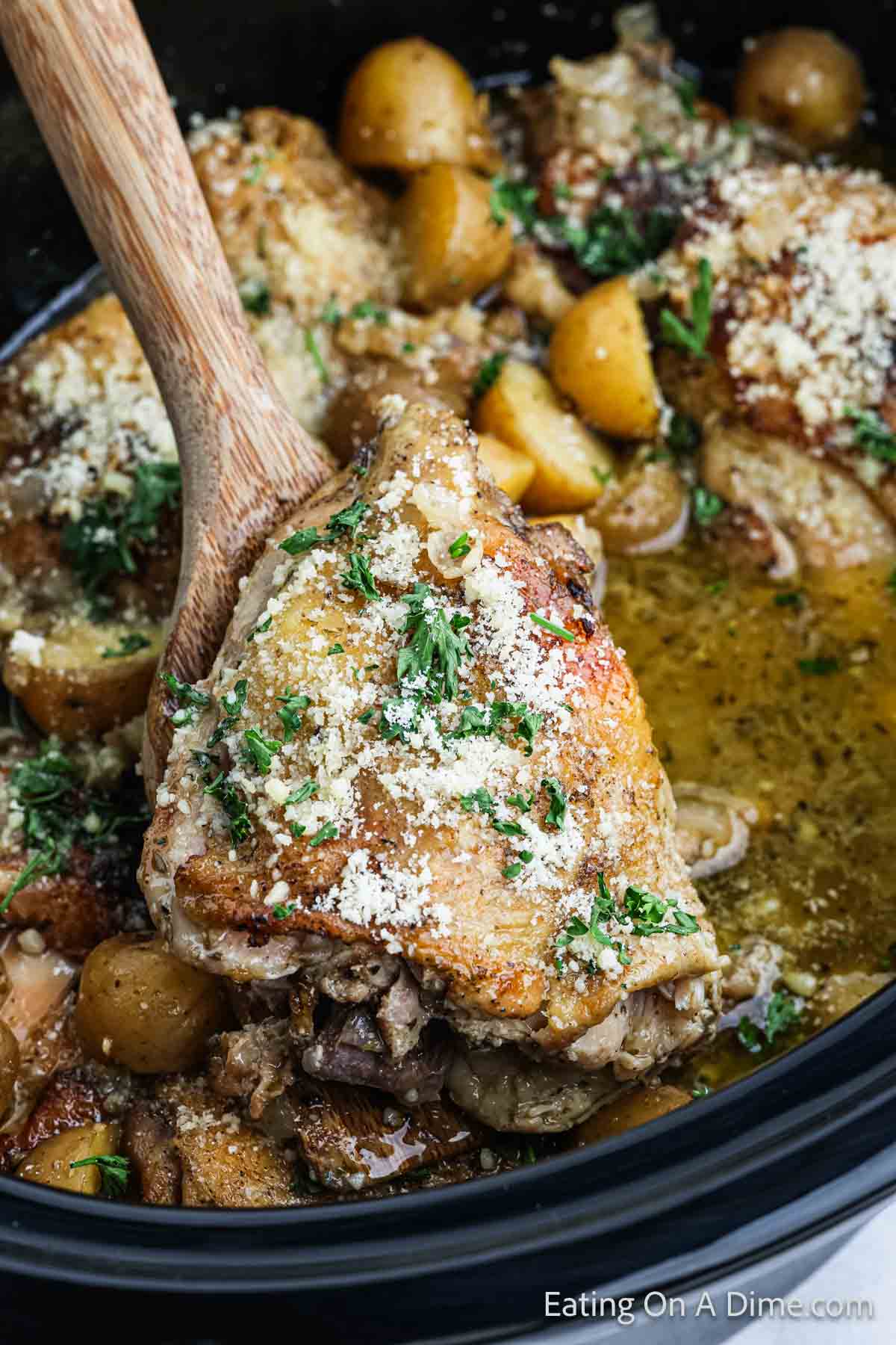 A wooden spoon lifts a piece of Slow Cooker Garlic Parmesan Chicken with a sprinkle of shredded cheese on top, surrounded by cooked baby potatoes and herbs in a rich broth. In the background is a black slow cooker with more of the dish. Text at the bottom reads "Eating On A Dime .com".