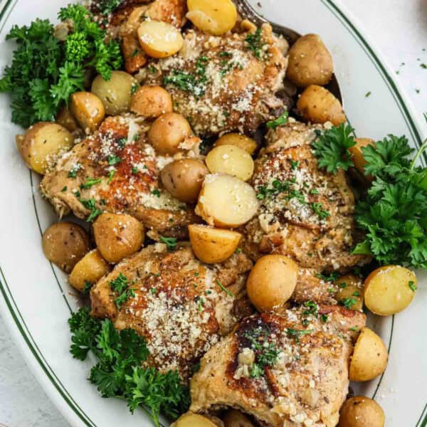 A white oval platter filled with seasoned roasted chicken thighs and baby potatoes, garnished with chopped parsley and grated cheese. The dish, reminiscent of Slow Cooker Garlic Parmesan Chicken, is vibrant and rustic, showcasing a golden-brown color and fresh herbs.