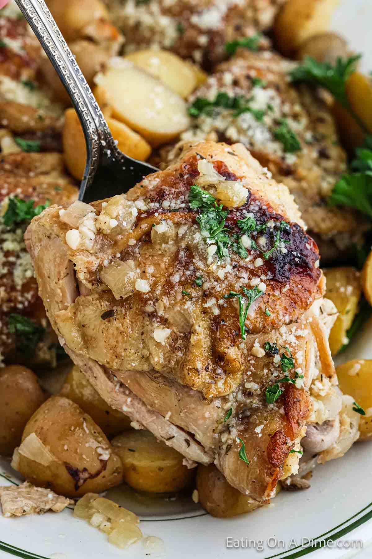 A close-up of a silver spoon holding a seasoned and cooked chicken thigh garnished with chopped parsley. It is surrounded by diced potatoes and additional pieces of Slow Cooker Garlic Parmesan Chicken, all sprinkled with grated cheese and herbs. The background shows more of the same dish.