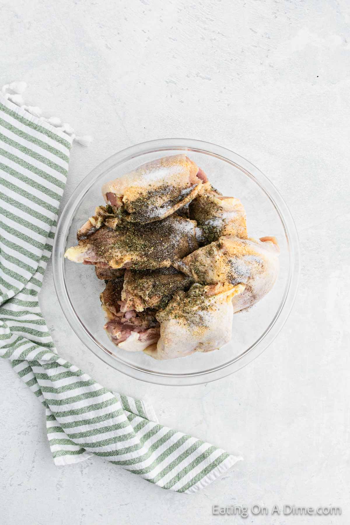 A clear glass bowl filled with seasoned Slow Cooker Garlic Parmesan Chicken pieces, placed on a light gray surface. A green and white striped kitchen towel is draped beside the bowl. "Eating on a Dime.com" is written in the bottom right corner of the image.