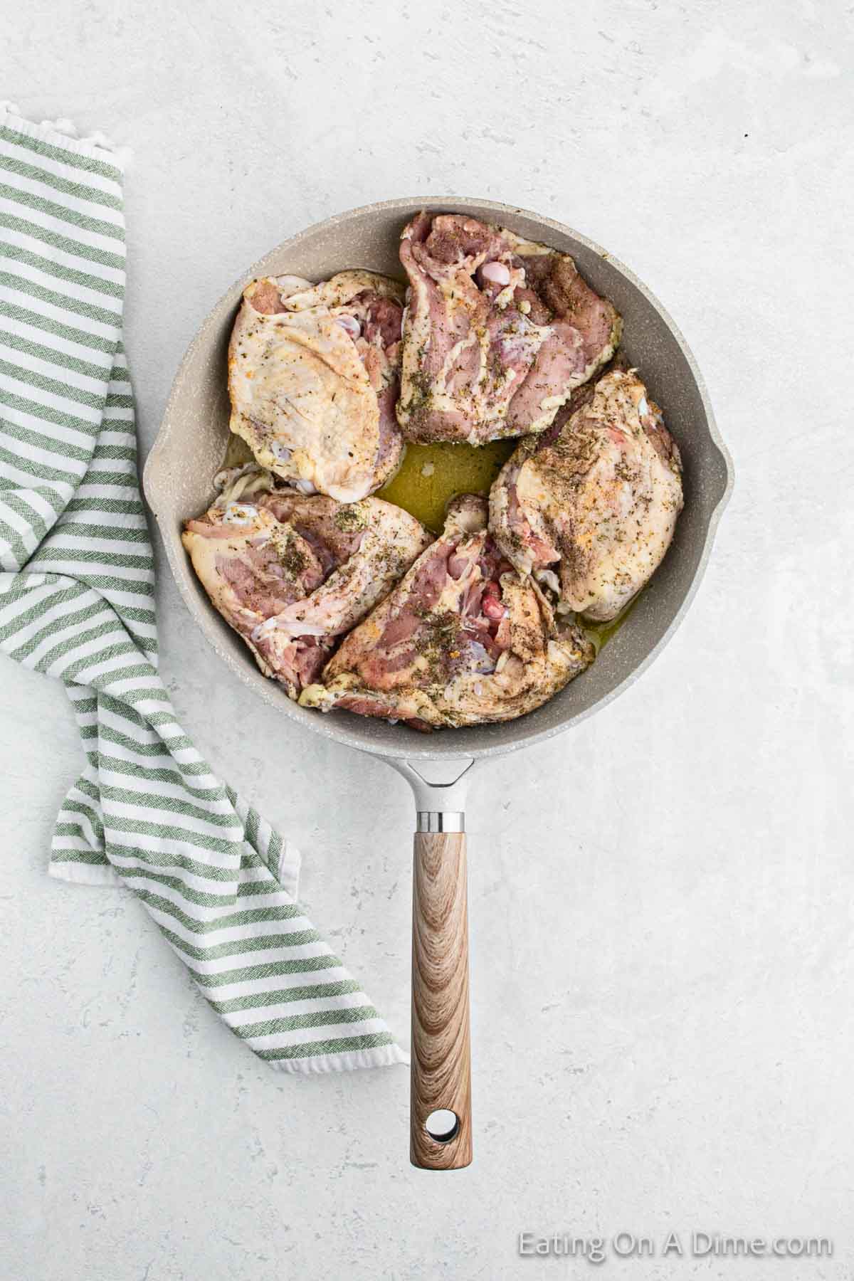 A frying pan with a wooden handle contains five pieces of seasoned raw chicken, reminiscent of Slow Cooker Garlic Parmesan Chicken. The pan rests on a light gray marble surface, with a green and white striped kitchen towel partially visible to the left.