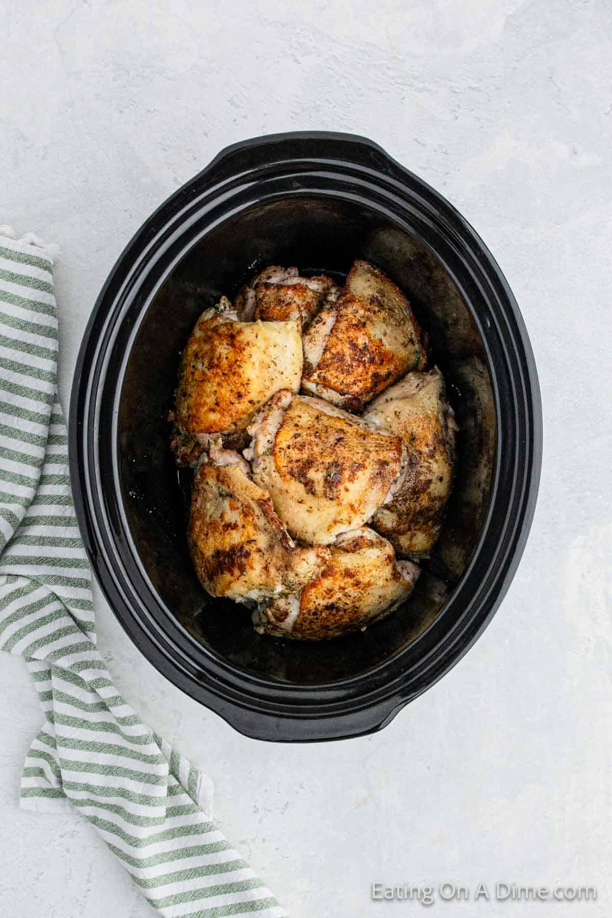 A black slow cooker filled with seasoned, cooked chicken thighs rests on a light gray countertop. The browned garlic Parmesan chicken thighs are arranged neatly inside the pot. Next to the slow cooker is a green and white striped kitchen towel.