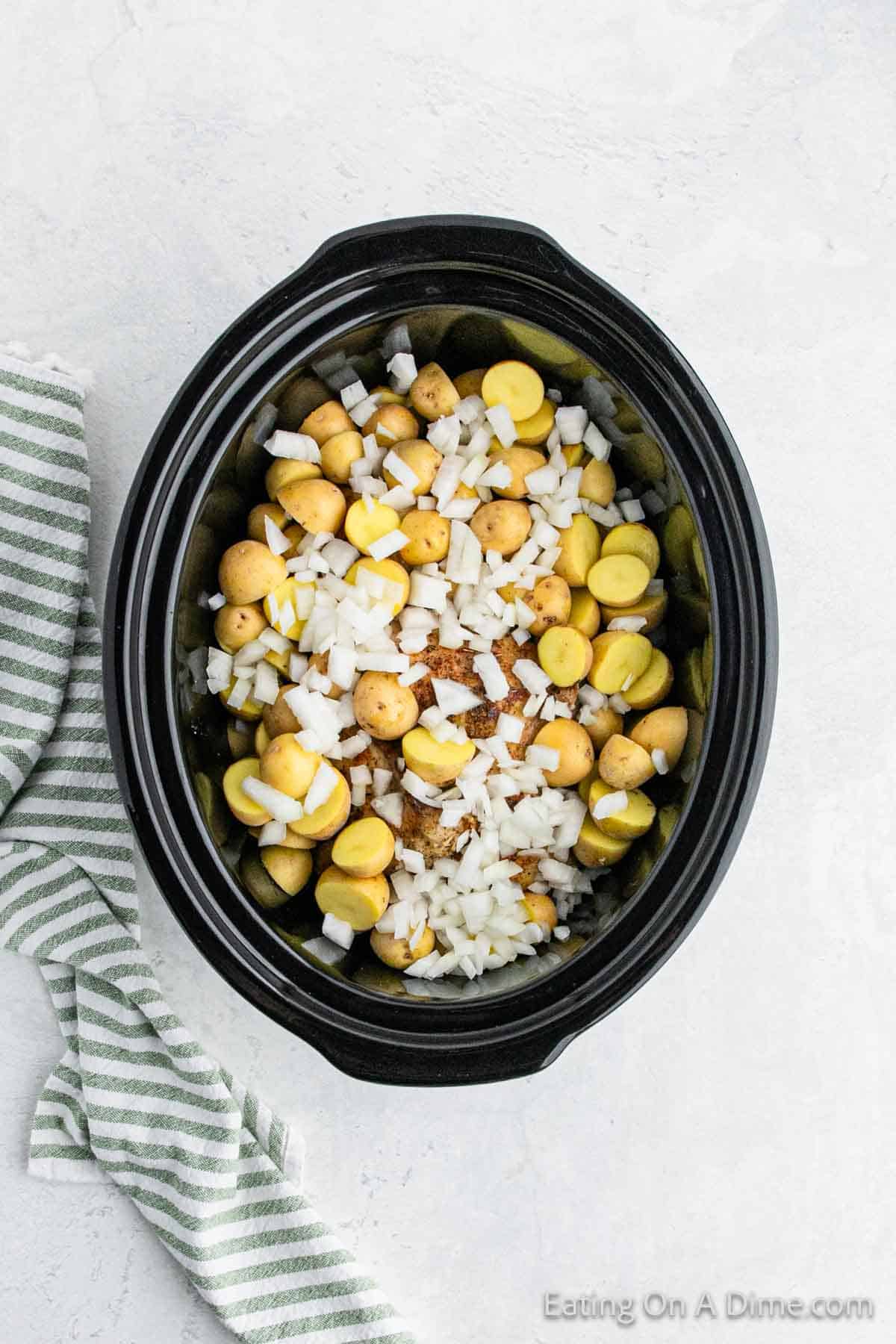 A slow cooker filled with halved baby potatoes, chopped onions, and seasoned for a hearty garlic Parmesan chicken dish. The black slow cooker sits on a light gray surface next to a green and white striped kitchen towel.