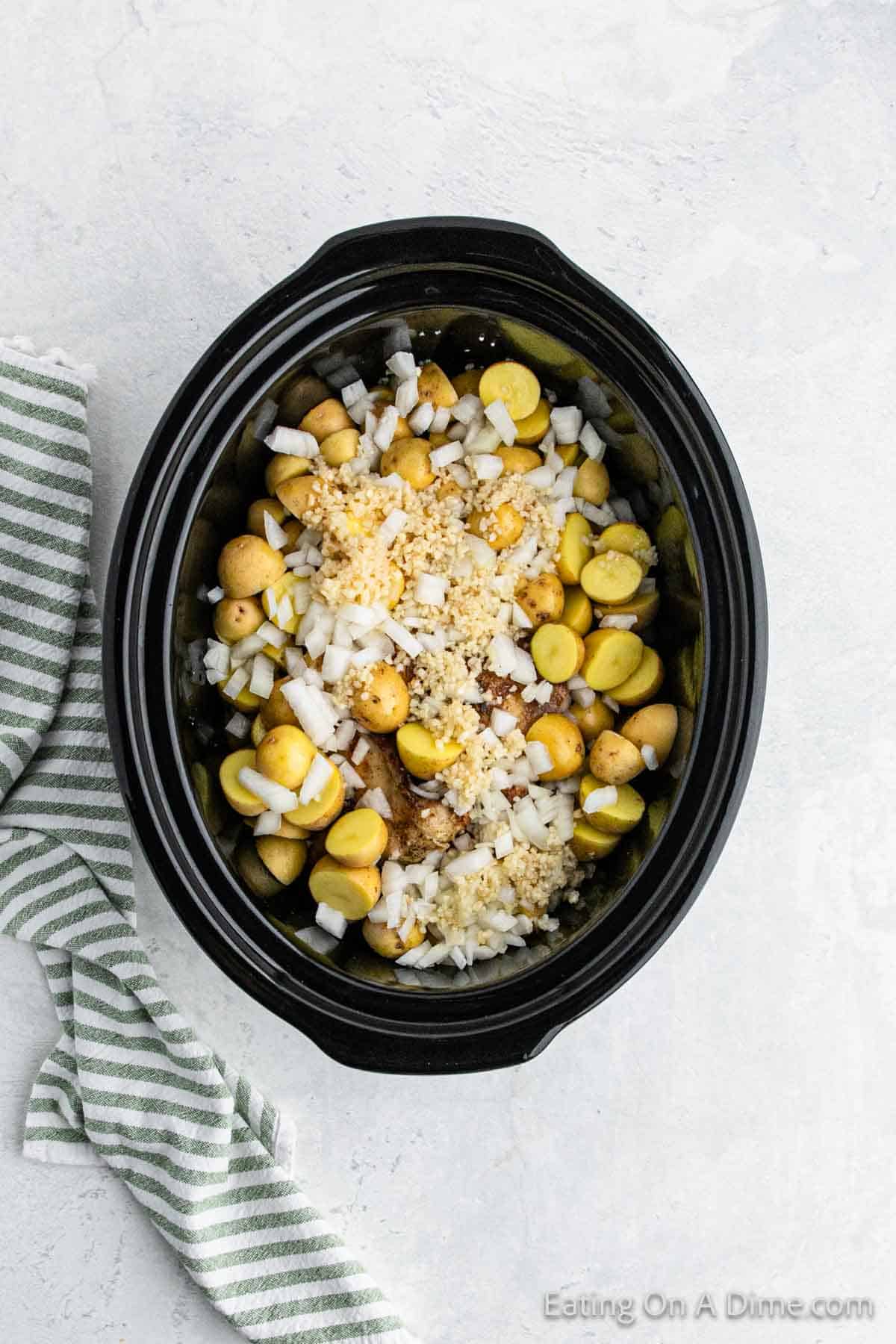 A black slow cooker brims with small yellow potatoes, chopped onions, and minced garlic on a light gray surface. To the left of it, a green and white striped kitchen towel is neatly folded. In the corner, "Eating On A Dime.com" teases a delicious recipe for Slow Cooker Garlic Parmesan Chicken.