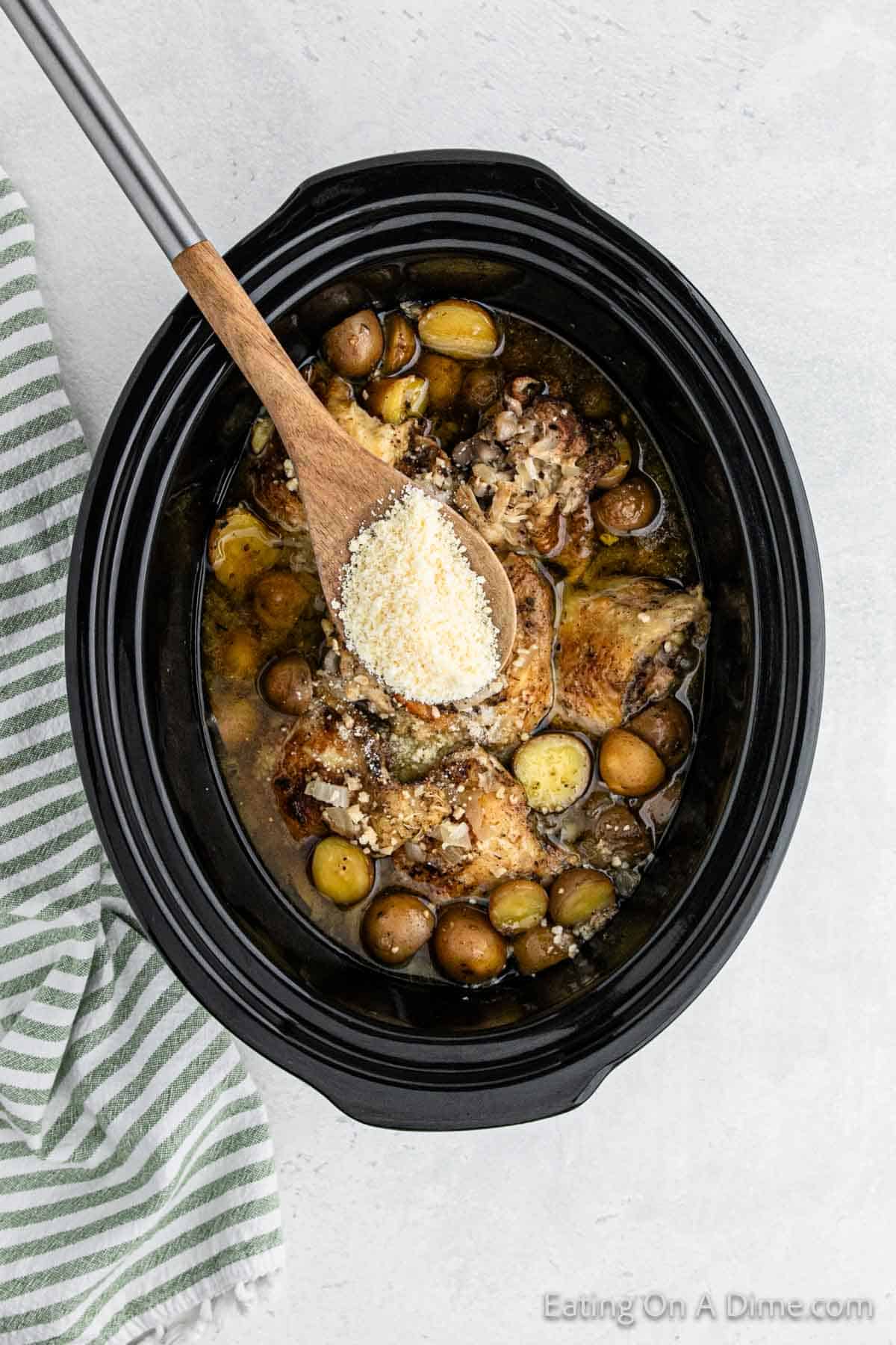 A slow cooker filled with Slow Cooker Garlic Parmesan Chicken, small potatoes, and a savory broth. A wooden spoon is sprinkling grated Parmesan cheese over the dish. A green and white striped towel is on the left side of the slow cooker. The image is branded with "Eating On A Dime.