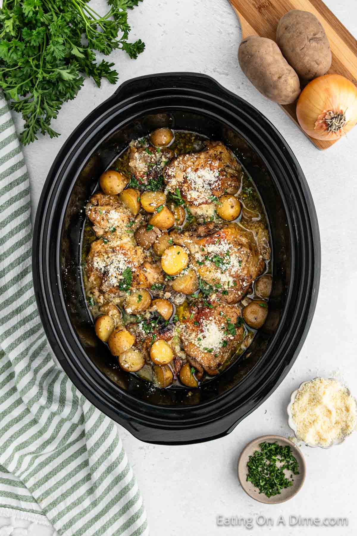 A slow cooker filled with Slow Cooker Garlic Parmesan Chicken, featuring seasoned and garnished chicken thighs and baby potatoes. The cooker is surrounded by parsley, potatoes, an onion on a cutting board, a bowl of grated cheese, chopped herbs in a small bowl, and a green striped cloth.