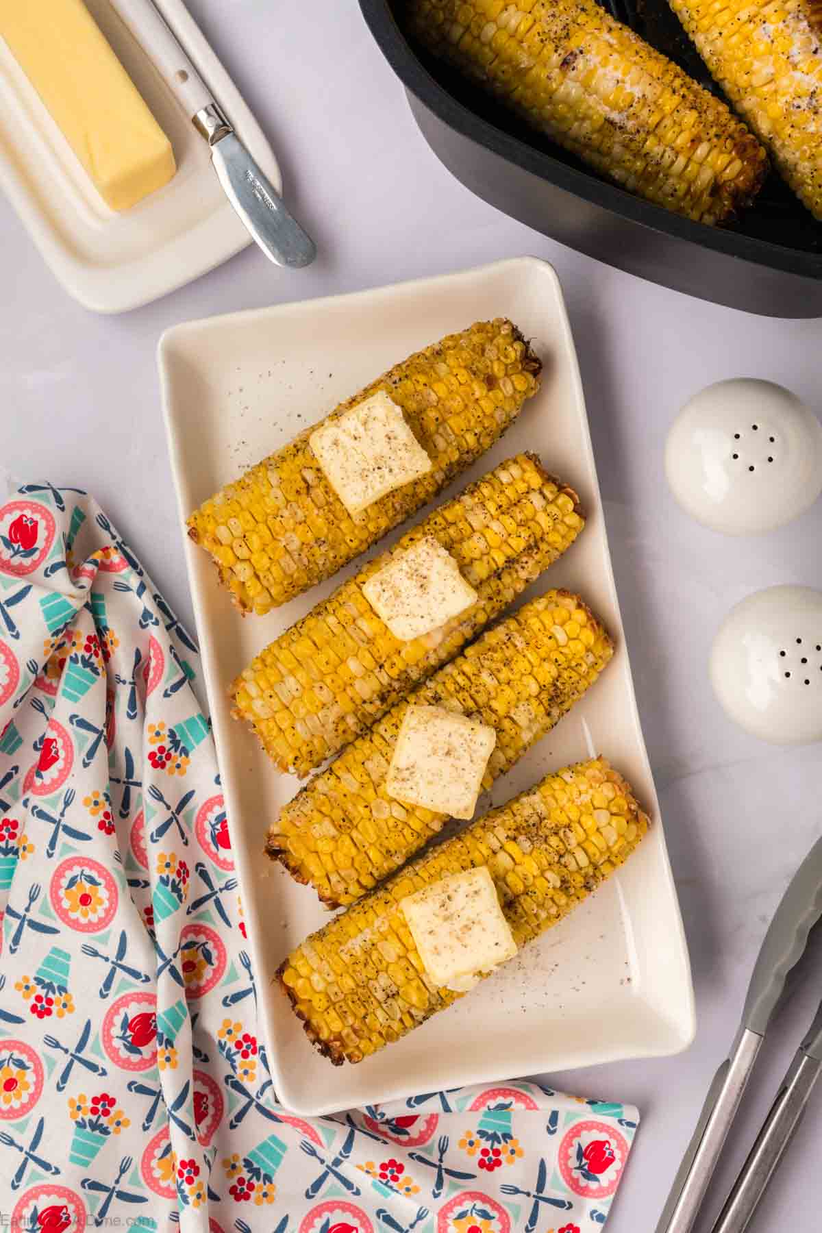 Four grilled corn on the cobs with butter pats and a sprinkle of seasoning are arranged on a white rectangular plate. A colorful patterned napkin, salt and pepper shakers, tongs, a butter dish with a knife, and additional air fryer corn on the cob in a black container are nearby.