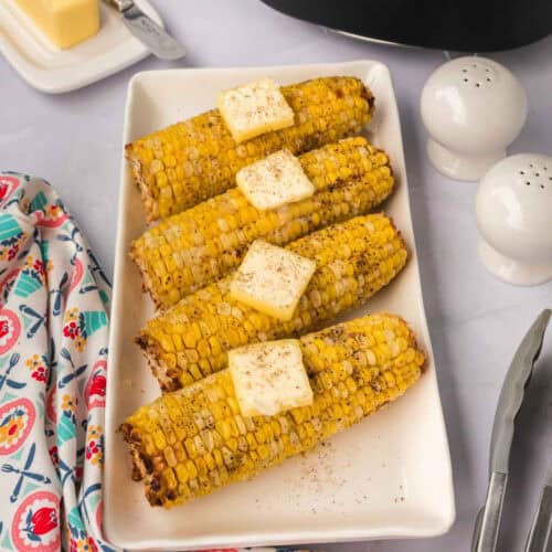 Four pieces of air fryer corn on the cob are arranged on a white rectangular platter. Each cob is topped with a square of butter and sprinkled with pepper. Salt and pepper shakers, a multicolored napkin, kitchen tongs, and a butter dish are nearby.