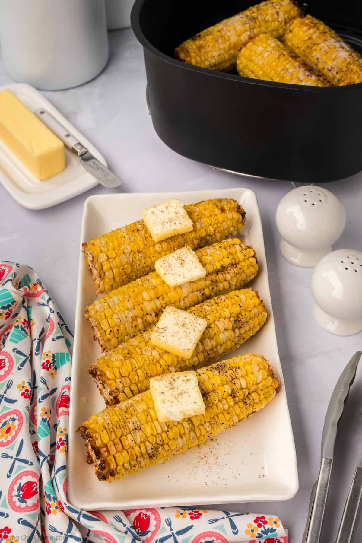 Air Fryer Corn on the Cob, charred to perfection and topped with butter and pepper, is arranged on a white rectangular plate. A pair of tongs, a floral-patterned napkin, salt and pepper shakers, and a butter dish are beside the plate. A pot with more corn is visible in the background.
