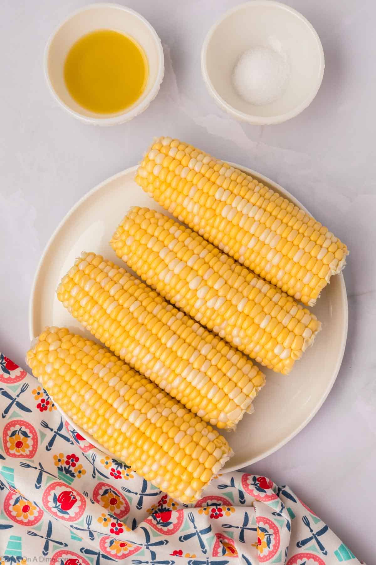 A plate of five corn on the cobs is placed on a table with a colorful floral-patterned cloth beside it. Near the plate, there are two small bowls, one containing oil and the other containing salt—perfect for seasoning your Air Fryer Corn on the Cob.