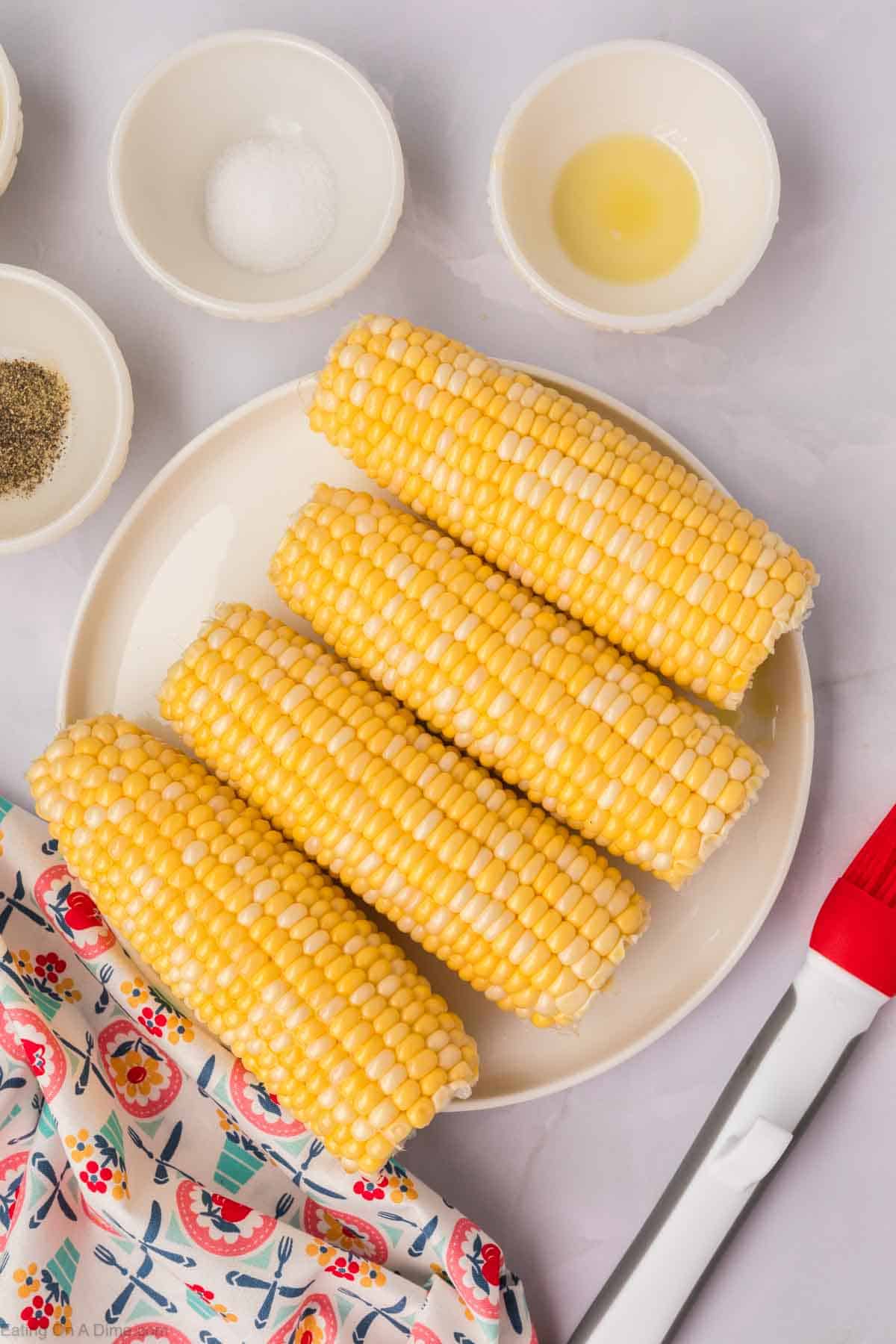 Four ears of Air Fryer Corn on the Cob are arranged on a round white plate. Surrounding the plate are small white bowls containing salt, pepper, butter, and oil. A decorative cloth and red brush are placed near the bottom of the image.