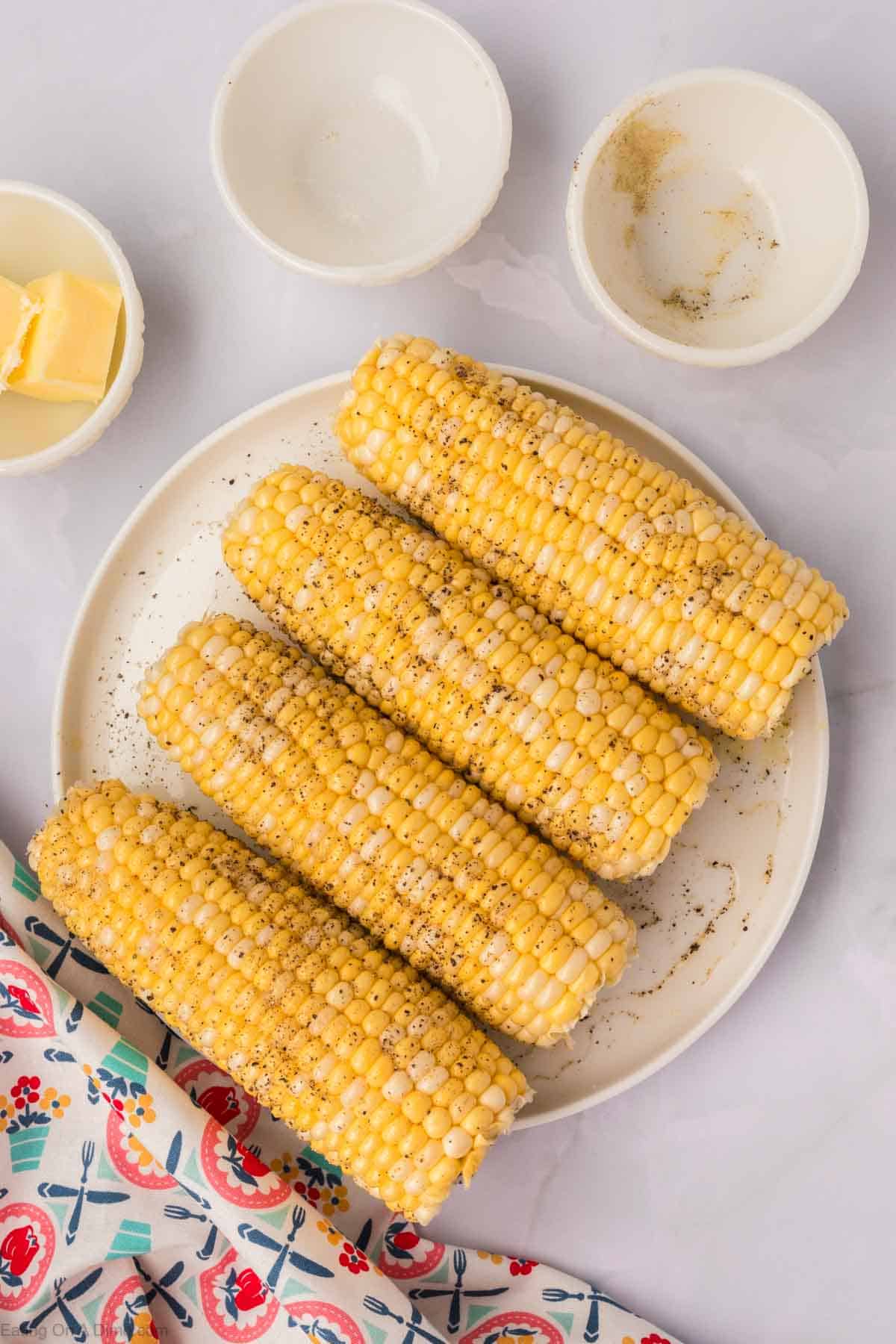 Four seasoned ears of corn, prepared as Air Fryer Corn on the Cob, are neatly arranged on a white plate with a sprinkle of pepper visible on their surface. Beside the plate, three small bowls sit, one containing a small piece of butter. A colorful, patterned cloth is partially visible in the foreground.
