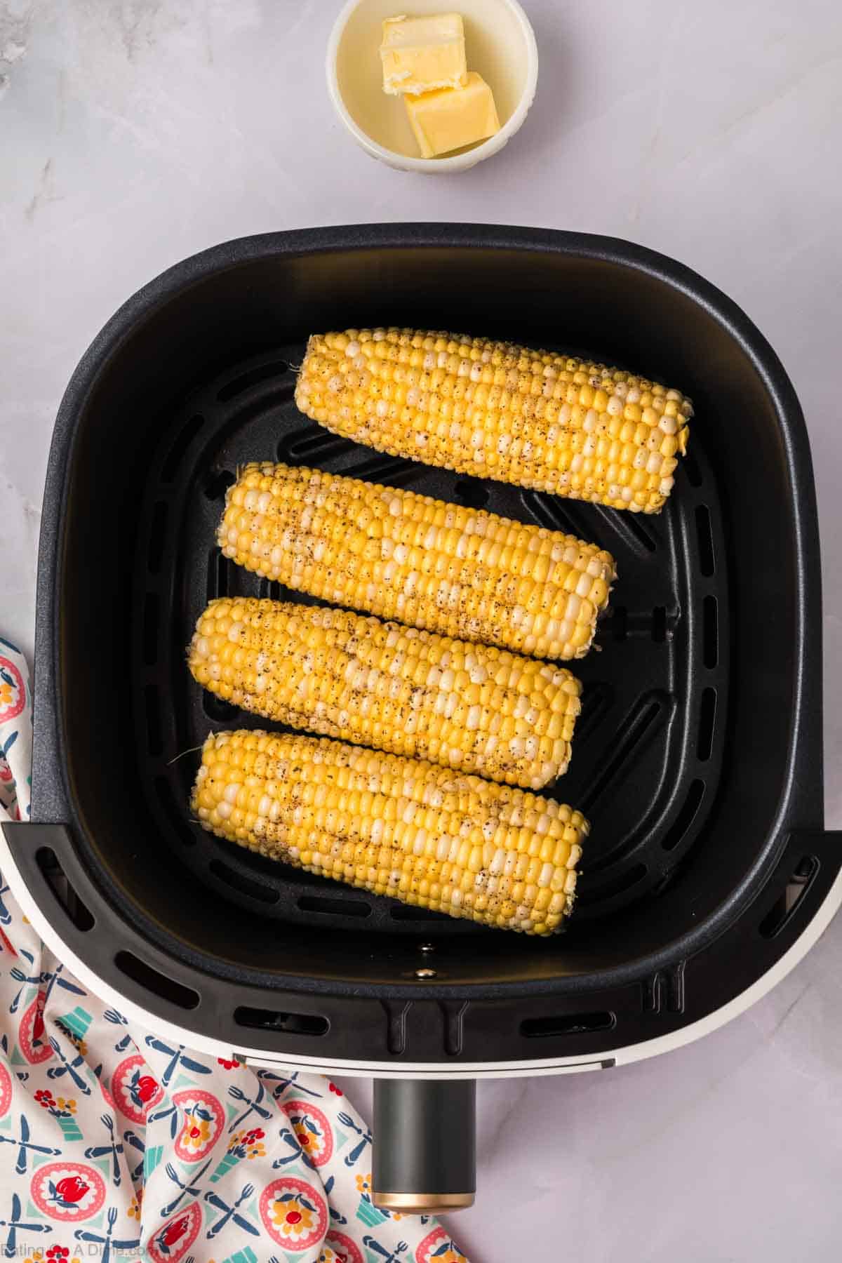 Four seasoned Air Fryer Corn on the Cob pieces stand vertically inside the basket. In the background, there's a bowl of butter, and to the left, a colorful patterned cloth napkin adds a pop of color.