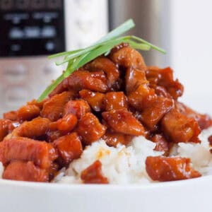 A bowl of white rice topped with glazed, diced meat garnished with green onions, reminiscent of an Instant Pot Honey Bourbon Chicken recipe. The background suggests the convenience of using an instant pot, and the dish looks savory and glossy.