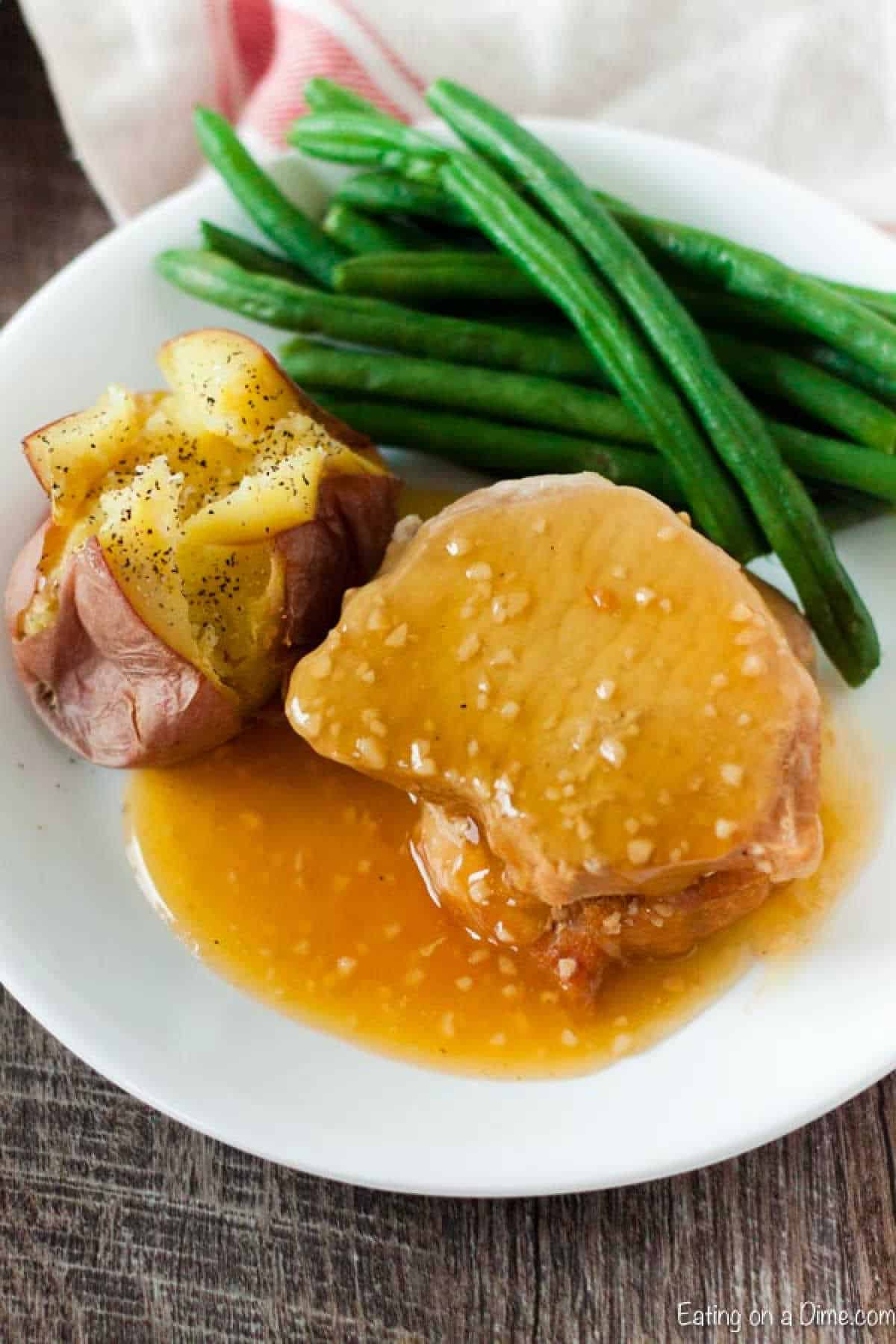 A white plate with green beans, a whole baked potato topped with cracked pepper, and succulent sliced pork chops drenched in golden honey garlic sauce, effortlessly prepared in an Instant Pot.