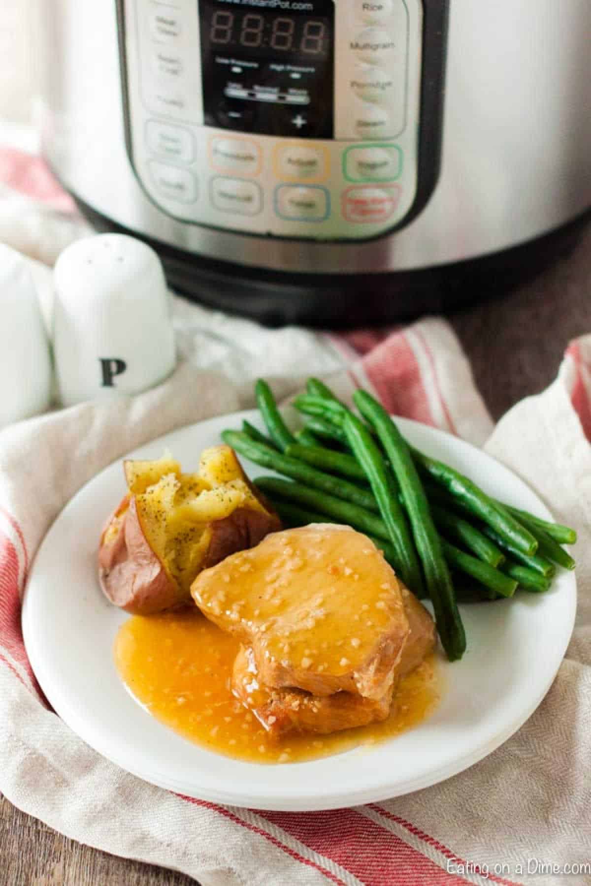 A plate featuring two honey garlic pork chops topped with sauce, a split baked potato sprinkled with butter and pepper, and a side of steamed green beans. In the background, an Instant Pot pressure cooker and salt and pepper shakers complete the cozy kitchen scene.