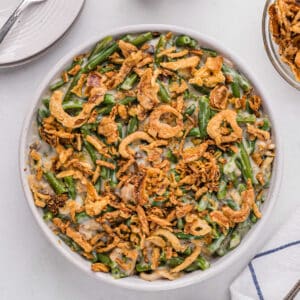 A round white dish filled with Instant Pot Green Bean Casserole topped with crispy fried onions sits on a white table. To the right, there's a partial view of a smaller bowl with more fried onions, and to the left, a plate with a fork. A white and blue napkin is in the bottom right.