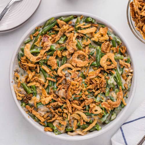 A round white dish filled with Instant Pot Green Bean Casserole topped with crispy fried onions sits on a white table. To the right, there's a partial view of a smaller bowl with more fried onions, and to the left, a plate with a fork. A white and blue napkin is in the bottom right.