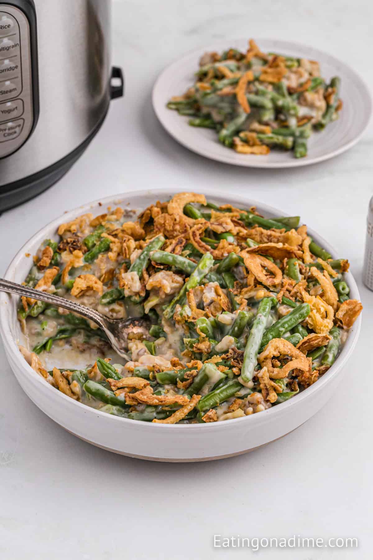 A serving dish filled with Instant Pot Green Bean Casserole sits on a countertop, with a spoon in the dish. A smaller plate with the casserole is in the background. Crispy fried onions top the green beans which are mixed with a creamy sauce, and an Instant Pot is partially visible.